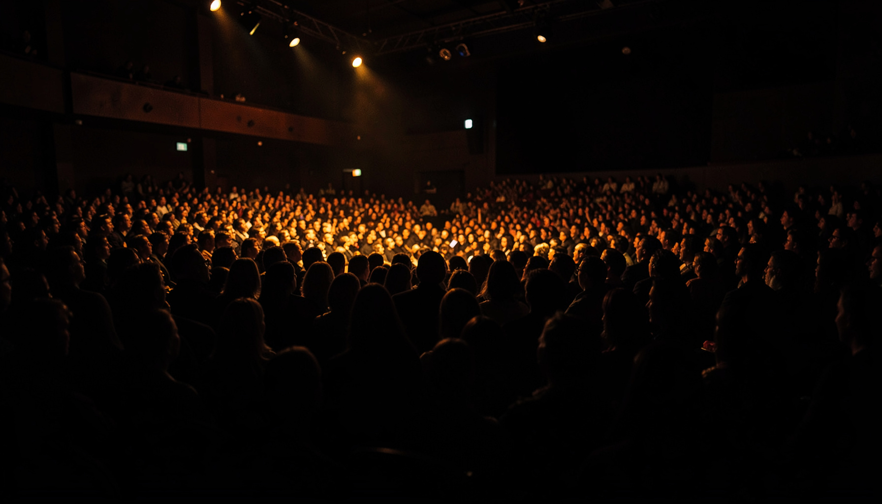 A dark, low-key photograph of a packed auditorium at a literary festival, warm amber stage lights cutting through deep shadow, silhouettes of audience members in the foreground, a sense of anticipation and community.