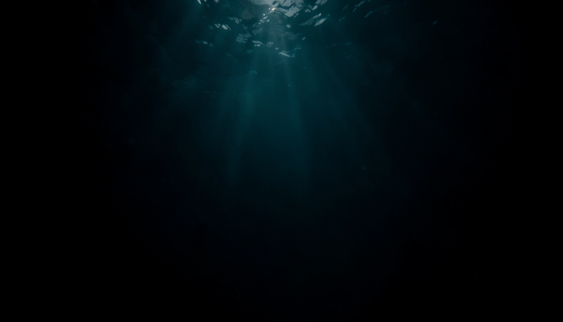A dark, low-key photograph of deep ocean water seen from below, with faint shafts of light filtering down through murky blue-black depths, evoking the vastness and mystery of space-like underwater environments.
